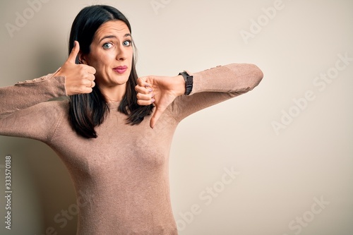 Young brunette woman with blue eyes wearing casual sweater over isolated white background Doing thumbs up and down, disagreement and agreement expression. Crazy conflict