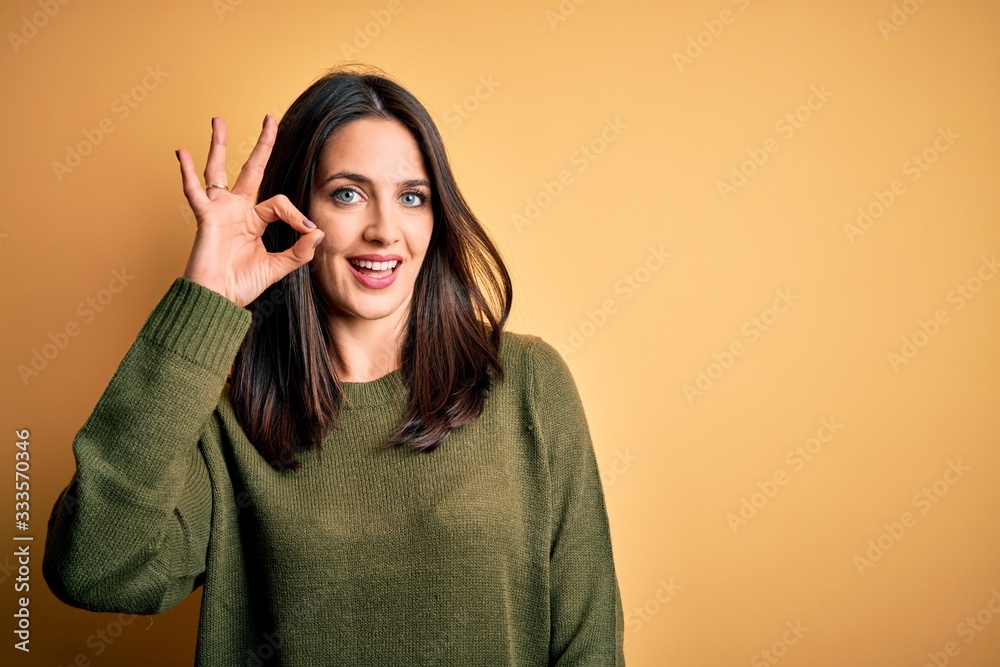 Φωτογραφία Young brunette woman with blue eyes wearing green casual sweater over yellow background smiling positive doing ok sign with hand and fingers