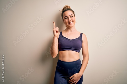 Young beautiful blonde sportswoman doing sport wearing sportswear over white background showing and pointing up with finger number one while smiling confident and happy.