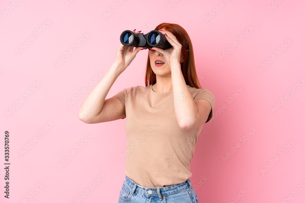 Young redhead woman over isolated pink background with black binoculars Young redhead woman over isolated pink background with black binoculars