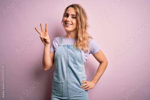 Young beautiful blonde woman wearing casual denim overalls standing over pink background showing and pointing up with fingers number three while smiling confident and happy. © Krakenimages.com