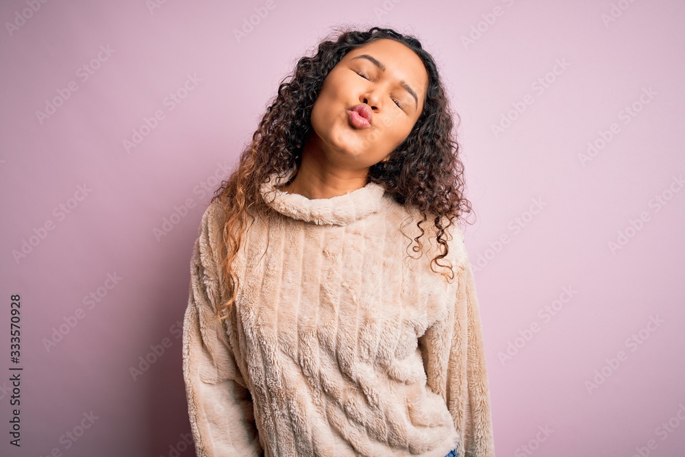 Young beautiful woman with curly hair wearing casual sweater standing over pink background looking at the camera blowing a kiss on air being lovely and sexy. Love expression. Young beautiful woman with curly hair wearing casual sweater standing over pink background looking at the camera blowing a kiss on air being lovely and sexy. Love expression.