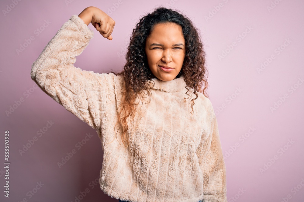 Young beautiful woman with curly hair wearing casual sweater standing over pink background Strong person showing arm muscle, confident and proud of power Young beautiful woman with curly hair wearing casual sweater standing over pink background Strong person showing arm muscle, confident and proud of power