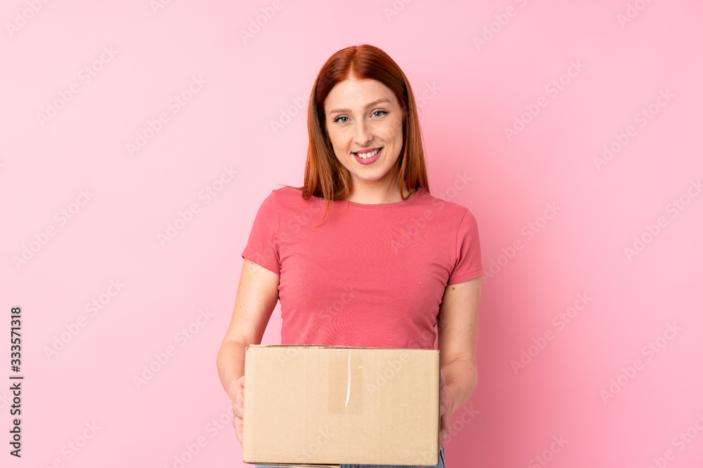 Young redhead woman over isolated pink background holding a box to move it to another site Young redhead woman over isolated pink background holding a box to move it to another site