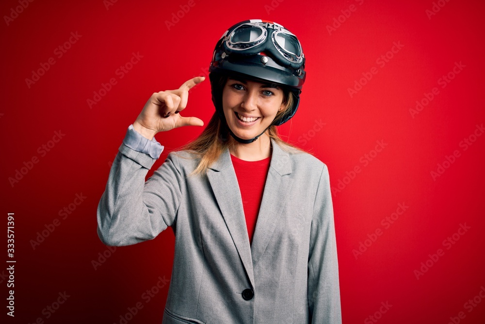 Young beautiful blonde motorcyclist woman wearing motorcycle helmet over red background smiling and confident gesturing with hand doing small size sign with fingers looking and the camera. Measure Young beautiful blonde motorcyclist woman wearing motorcycle helmet over red background smiling and confident gesturing with hand doing small size sign with fingers looking and the camera. Measure