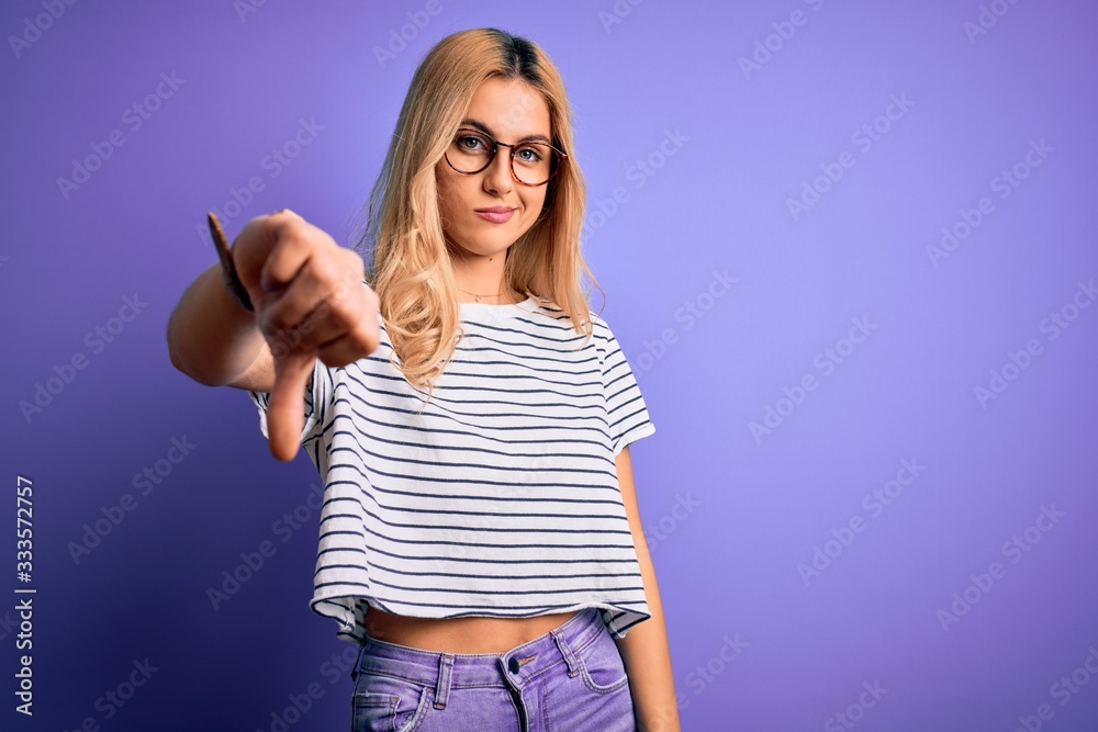 Young beautiful blonde woman wearing striped t-shirt and glasses over purple background looking unhappy and angry showing rejection and negative with thumbs down gesture. Bad expression. Young beautiful blonde woman wearing striped t-shirt and glasses over purple background looking unhappy and angry showing rejection and negative with thumbs down gesture. Bad expression.
