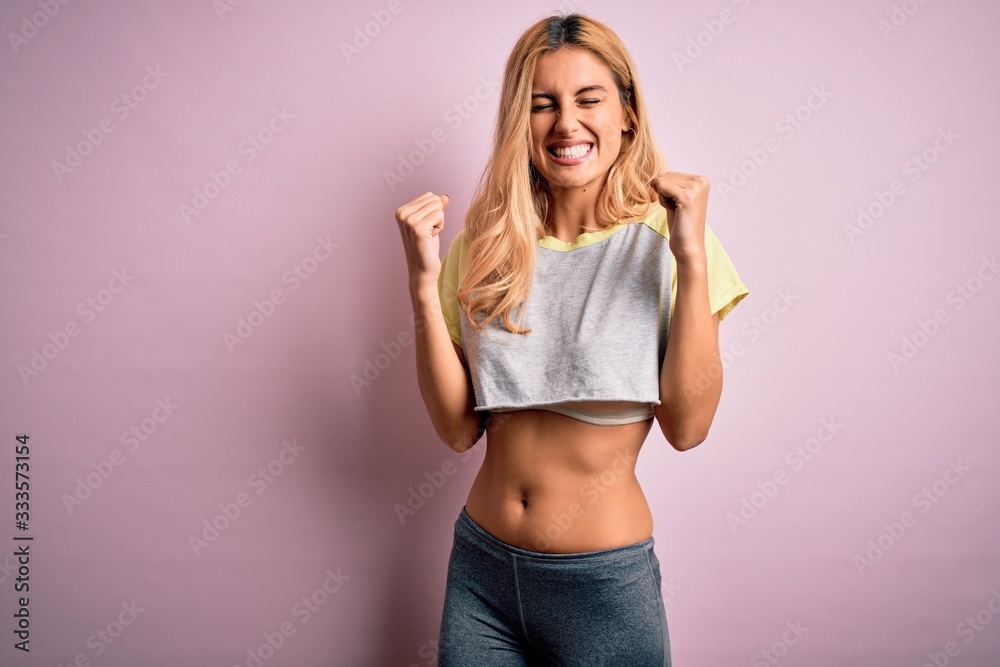 Young beautiful blonde sportswoman doing sport wearing sportswear over pink background very happy and excited doing winner gesture with arms raised, smiling and screaming for success. Celebration 