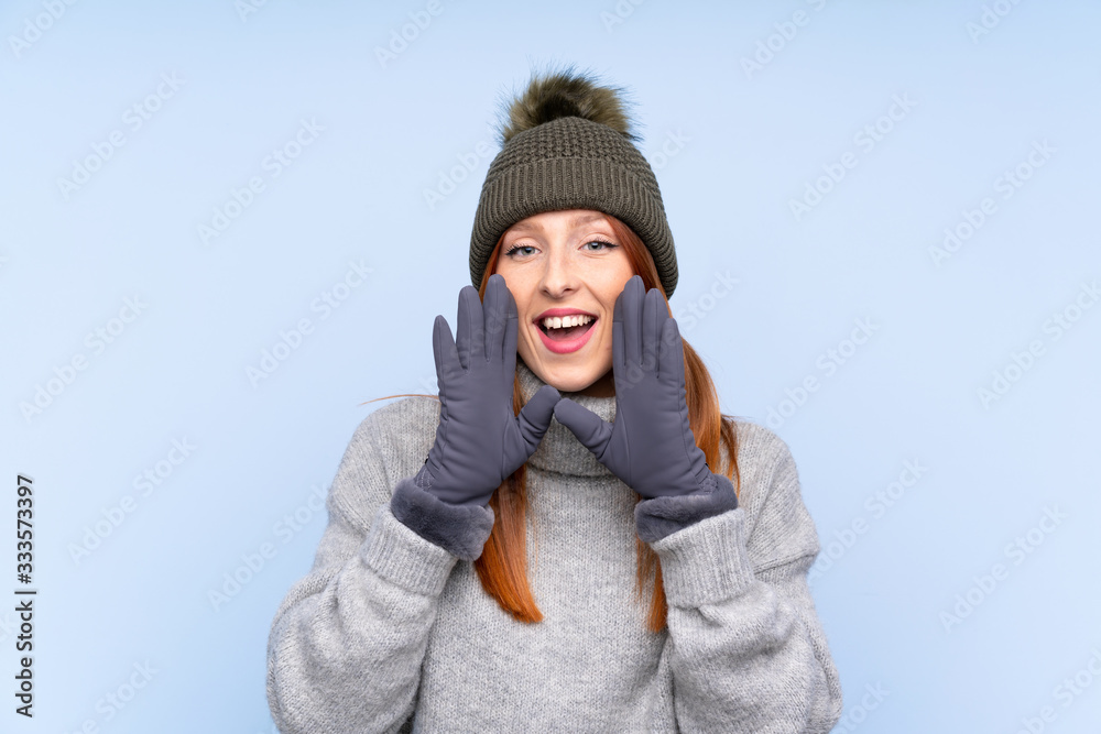 Young redhead Russian woman with winter hat over isolated blue background shouting with mouth wide open Young redhead Russian woman with winter hat over isolated blue background shouting with mouth wide open
