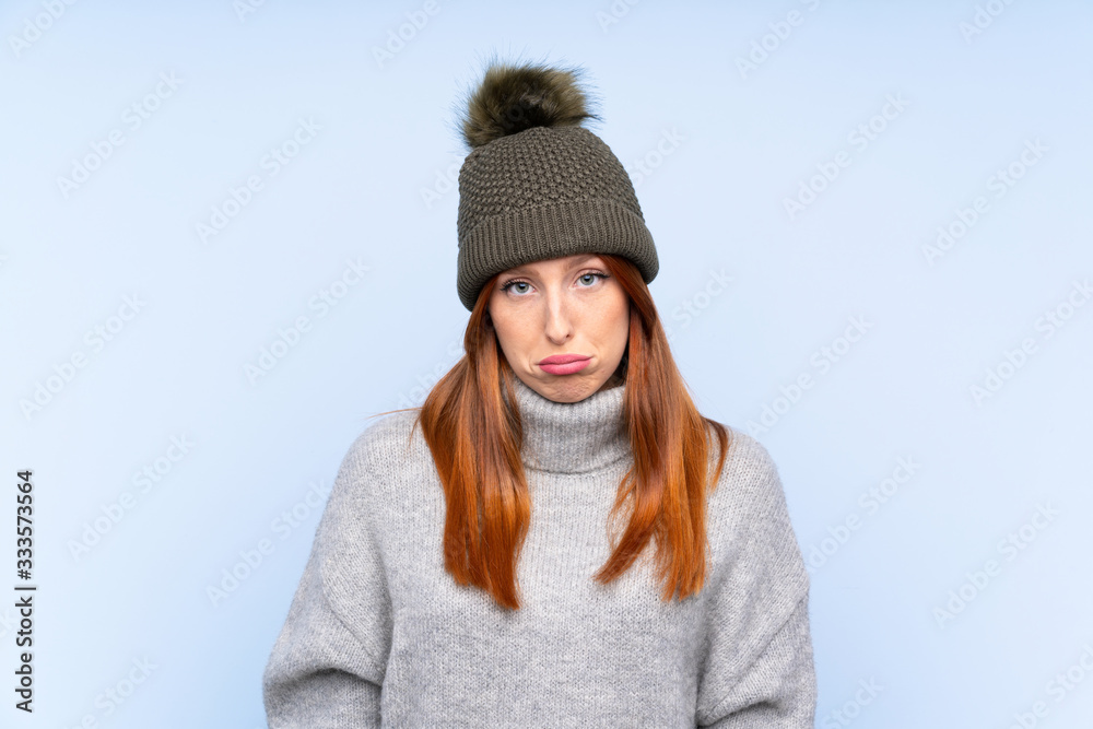 Young redhead Russian woman with winter hat over isolated blue background sad Young redhead Russian woman with winter hat over isolated blue background sad