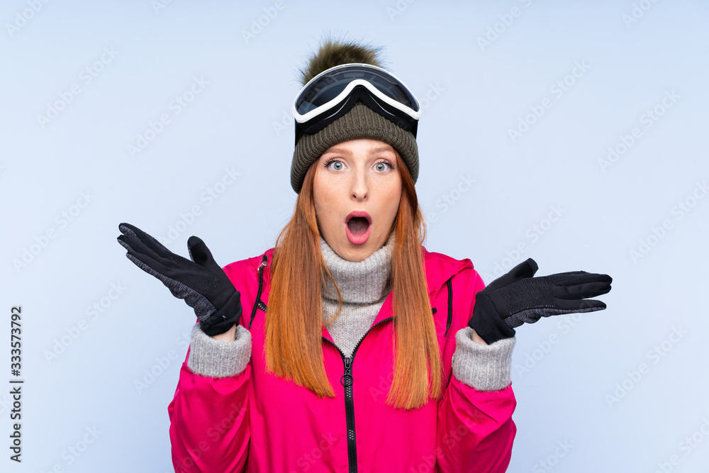 Skier redhead woman with snowboarding glasses over isolated blue wall with surprise facial expression Skier redhead woman with snowboarding glasses over isolated blue wall with surprise facial expression