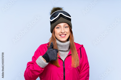 Skier redhead woman with snowboarding glasses over isolated blue wall giving a thumbs up gesture