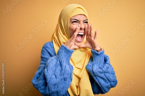Young beautiful brunette muslim woman wearing arab hijab over isolated yellow background Shouting angry out loud with hands over mouth