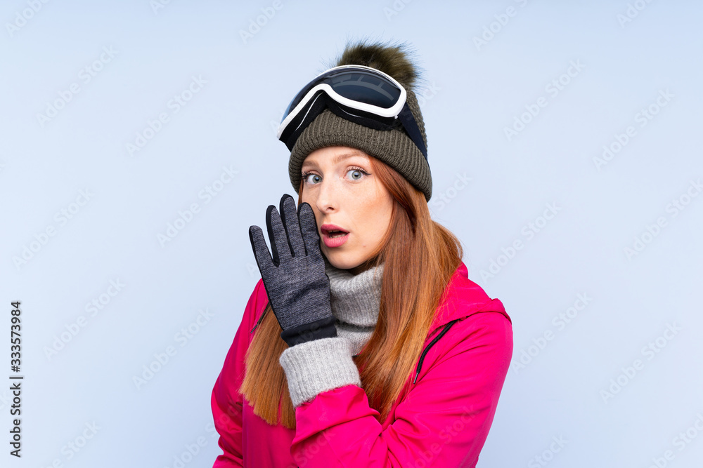 Skier redhead woman with snowboarding glasses over isolated blue wall whispering something