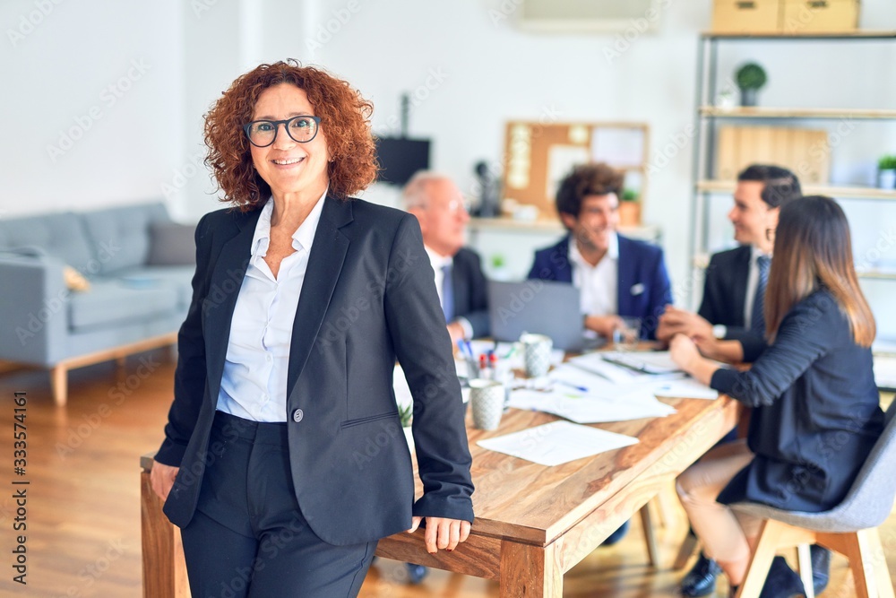 Group of business workers smiling happy and confident working together in a meeting. One of them, standing with smile on face looking at camera at the office. Group of business workers smiling happy and confident working together in a meeting. One of them, standing with smile on face looking at camera at the office.