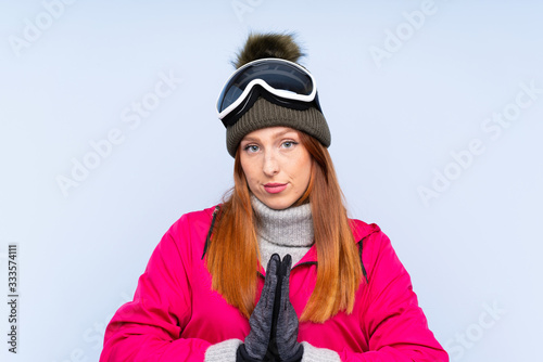 Skier redhead woman with snowboarding glasses over isolated blue wall pleading © luismolinero