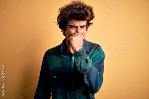 Young handsome man wearing casual shirt standing over isolated yellow background smelling something stinky and disgusting, intolerable smell, holding breath with fingers on nose. Bad smell © Krakenimages.com