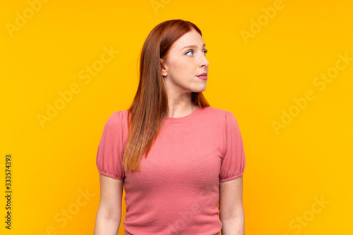 Young redhead woman over isolated yellow background looking side