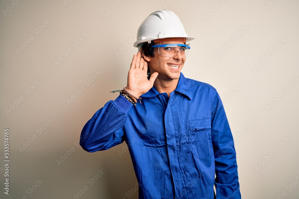 Young constructor man wearing uniform and security helmet over isolated white background smiling with hand over ear listening an hearing to rumor or gossip. Deafness concept.