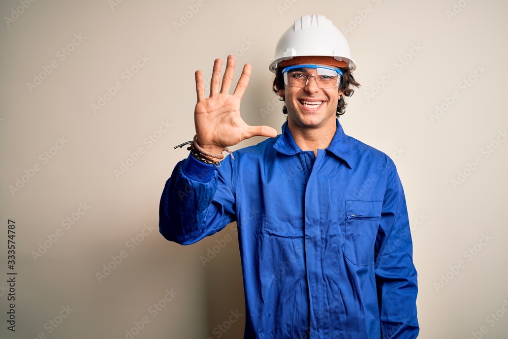 Young constructor man wearing uniform and security helmet over isolated white background showing and pointing up with fingers number five while smiling confident and happy.
