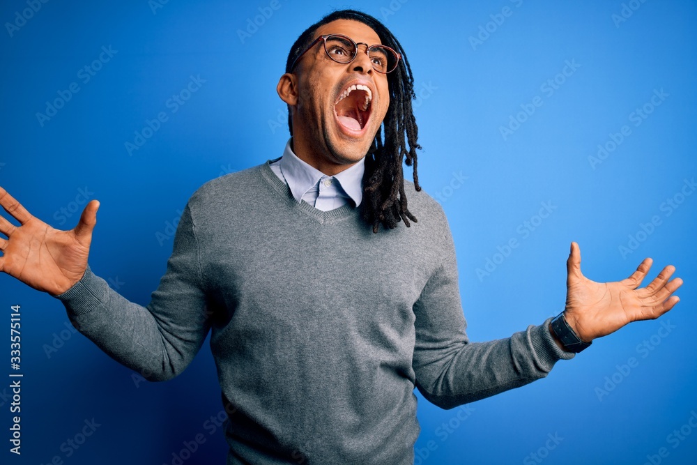 Fototapeta premium Young handsome african american man with dreadlocks wearing casual sweater and glasses crazy and mad shouting and yelling with aggressive expression and arms raised. Frustration concept.