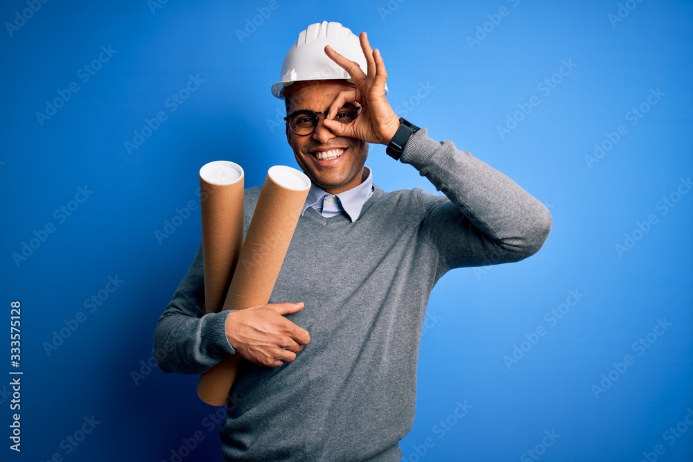 Naklejka premium Young african american afro architect man wearing safety helmet holding bluprints with happy face smiling doing ok sign with hand on eye looking through fingers
