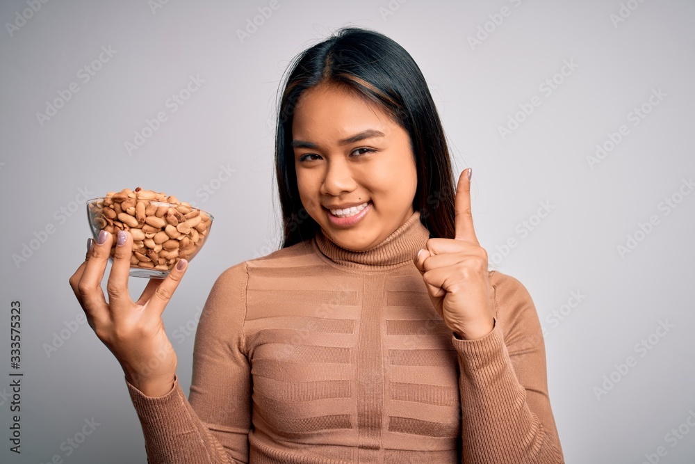 Young asian girl holding bowl of healthy peanuts eating as snack for diet over white background surprised with an idea or question pointing finger with happy face, number one Young asian girl holding bowl of healthy peanuts eating as snack for diet over white background surprised with an idea or question pointing finger with happy face, number one