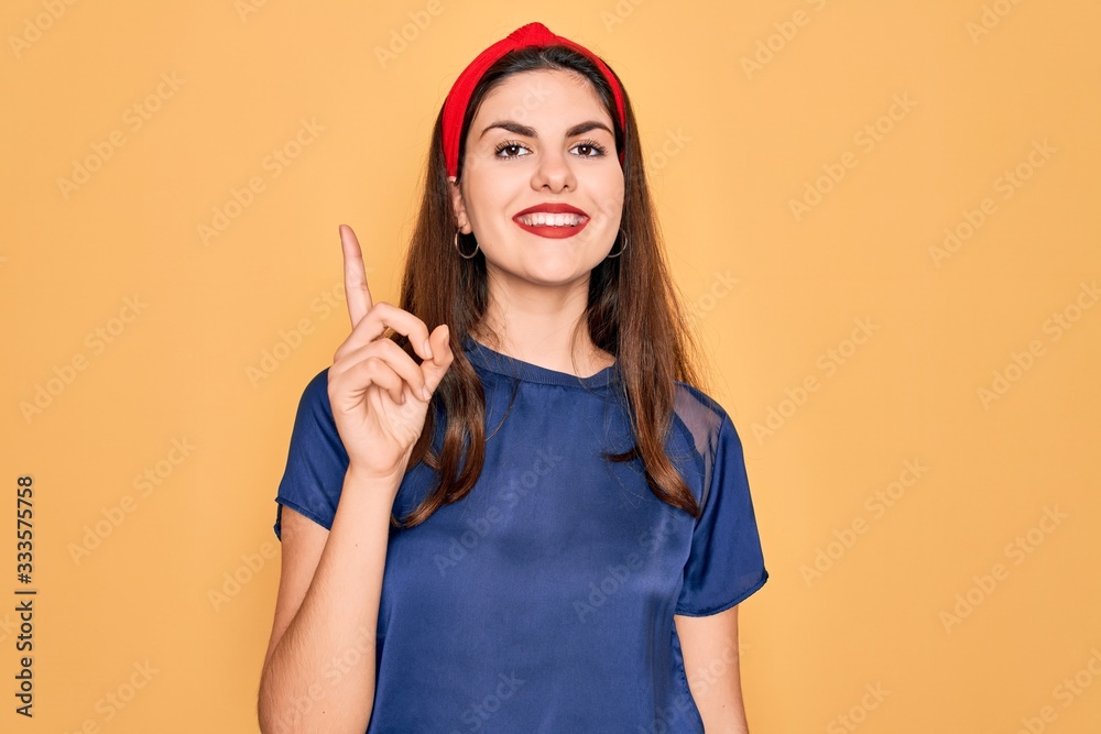 Young beautiful brunette woman wearing red lips over yellow background showing and pointing up with finger number one while smiling confident and happy. Young beautiful brunette woman wearing red lips over yellow background showing and pointing up with finger number one while smiling confident and happy.