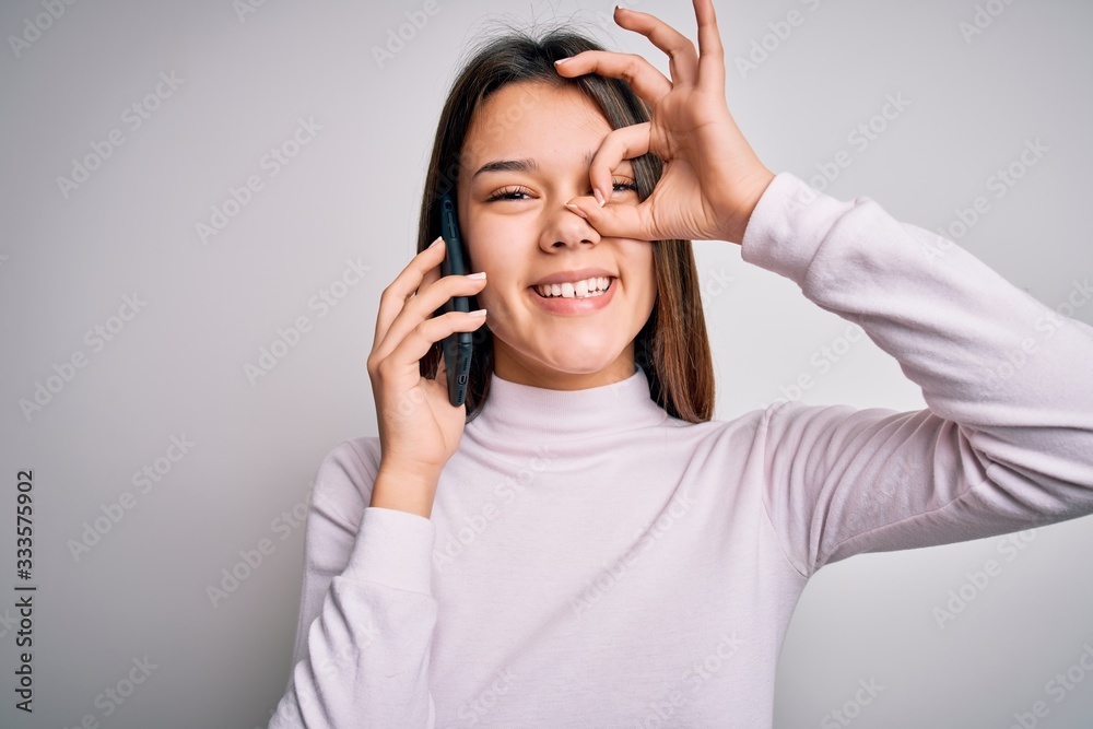 Beautiful brunette girl having conversation talking on the smartphone over white background with happy face smiling doing ok sign with hand on eye looking through fingers Beautiful brunette girl having conversation talking on the smartphone over white background with happy face smiling doing ok sign with hand on eye looking through fingers