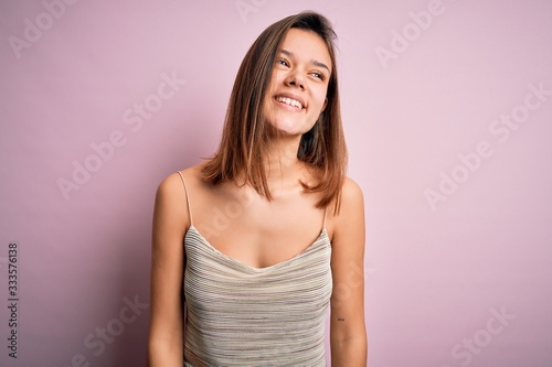 Young beautiful brunette girl wearing casual striped t-shirt over isolated pink background looking away to side with smile on face, natural expression. Laughing confident.