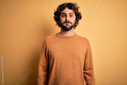 Young handsome man with beard wearing casual sweater standing over yellow background puffing cheeks with funny face. Mouth inflated with air, crazy expression.