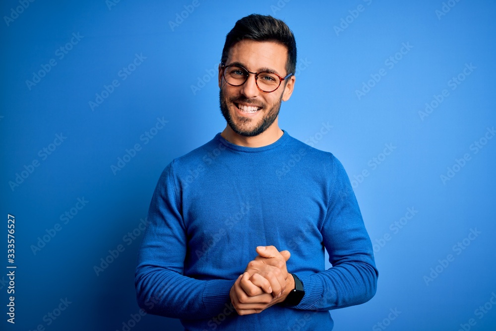 Obraz premium Young handsome man with beard wearing casual sweater and glasses over blue background with hands together and crossed fingers smiling relaxed and cheerful. Success and optimistic