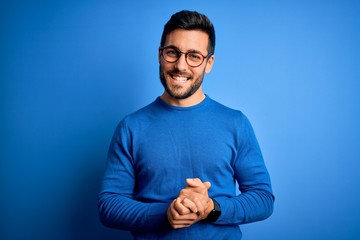 Young handsome man with beard wearing casual sweater and glasses over blue background with hands together and crossed fingers smiling relaxed and cheerful. Success and optimistic