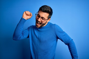 Young handsome man with beard wearing casual sweater and glasses over blue background Dancing happy and cheerful, smiling moving casual and confident listening to music