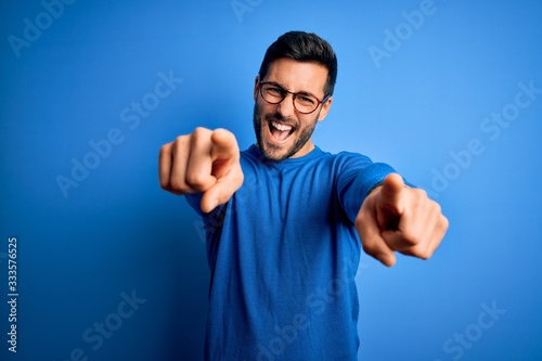 Young handsome man with beard wearing casual sweater and glasses over blue background pointing to you and the camera with fingers, smiling positive and cheerful