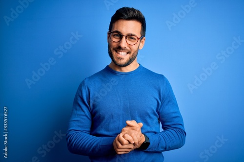Foto Young handsome man with beard wearing casual sweater and glasses over blue background with hands together and crossed fingers smiling relaxed and cheerful