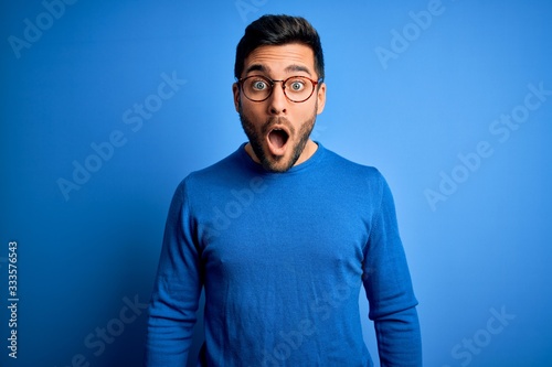 Young handsome man with beard wearing casual sweater and glasses over blue background afraid and shocked with surprise and amazed expression, fear and excited face.