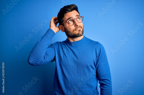 Young handsome man with beard wearing casual sweater and glasses over blue background confuse and wondering about question. Uncertain with doubt, thinking with hand on head. Pensive concept. photo