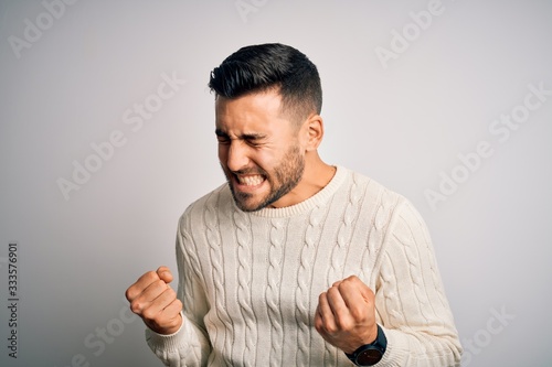 Young handsome man wearing casual sweater standing over isolated white background very happy and excited doing winner gesture with arms raised, smiling and screaming for success. Celebration concept.