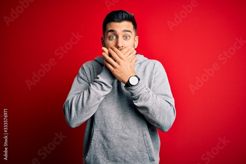 Young handsome sportsman wearing sweatshirt standing over isolated red background shocked covering mouth with hands for mistake. Secret concept.