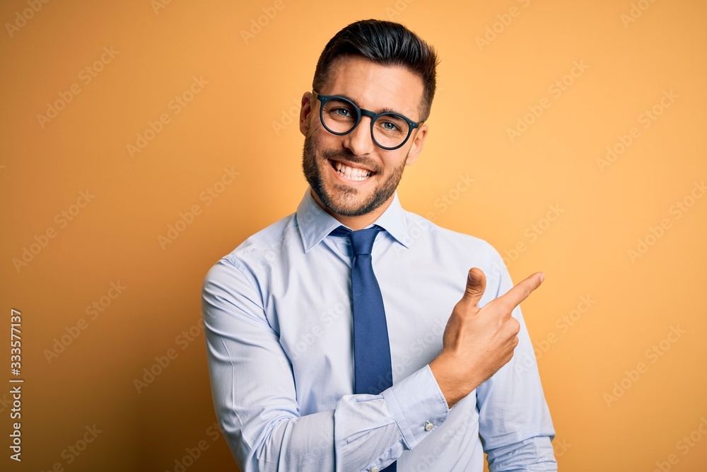 Young handsome businessman wearing tie and glasses standing over yellow background cheerful with a smile on face pointing with hand and finger up to the side with happy and natural expression Young handsome businessman wearing tie and glasses standing over yellow background cheerful with a smile on face pointing with hand and finger up to the side with happy and natural expression