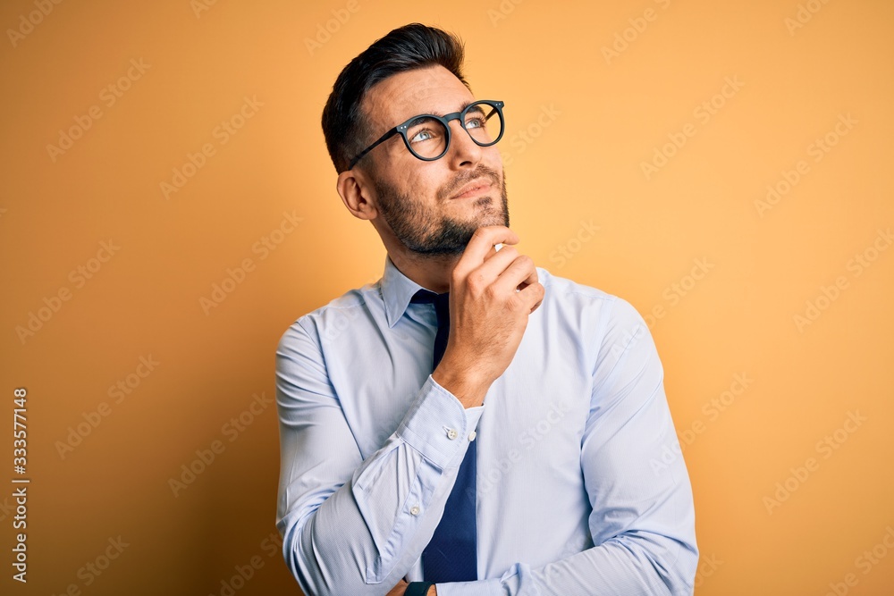 Young handsome businessman wearing tie and glasses standing over yellow background with hand on chin thinking about question, pensive expression. Smiling and thoughtful face. Doubt concept. Young handsome businessman wearing tie and glasses standing over yellow background with hand on chin thinking about question, pensive expression. Smiling and thoughtful face. Doubt concept.