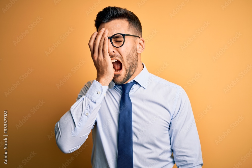 Young handsome businessman wearing tie and glasses standing over yellow background Yawning tired covering half face, eye and mouth with hand. Face hurts in pain. Young handsome businessman wearing tie and glasses standing over yellow background Yawning tired covering half face, eye and mouth with hand. Face hurts in pain.