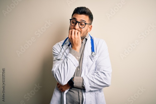 Young doctor man wearing glasses, medical white robe and stethoscope over isolated background looking stressed and nervous with hands on mouth biting nails. Anxiety problem.