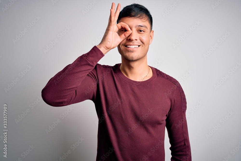 Young handsome hispanic man wearing casual shirt standing over white isolated background doing ok gesture with hand smiling, eye looking through fingers with happy face. Young handsome hispanic man wearing casual shirt standing over white isolated background doing ok gesture with hand smiling, eye looking through fingers with happy face.
