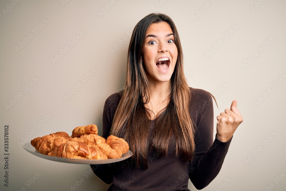 Young beautiful girl holding plate with sweet croissants for breakfast over white background screaming proud and celebrating victory and success very excited, cheering emotion Young beautiful girl holding plate with sweet croissants for breakfast over white background screaming proud and celebrating victory and success very excited, cheering emotion