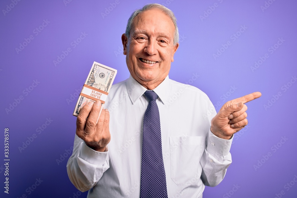 Senior grey haired man holding bunch of fifty dollars banknotes over purple background very happy pointing with hand and finger to the side Senior grey haired man holding bunch of fifty dollars banknotes over purple background very happy pointing with hand and finger to the side