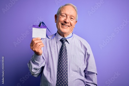 Senior grey haired business man holding identification tag over purple background with a happy face standing and smiling with a confident smile showing teeth