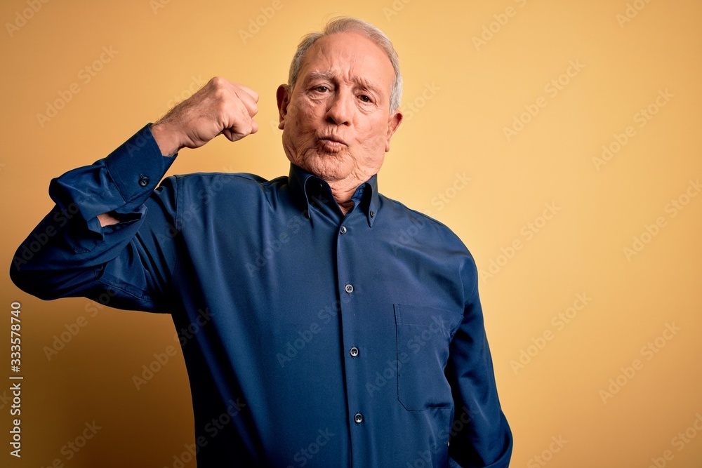 Grey haired senior man wearing casual blue shirt standing over yellow background Strong person showing arm muscle, confident and proud of power Grey haired senior man wearing casual blue shirt standing over yellow background Strong person showing arm muscle, confident and proud of power