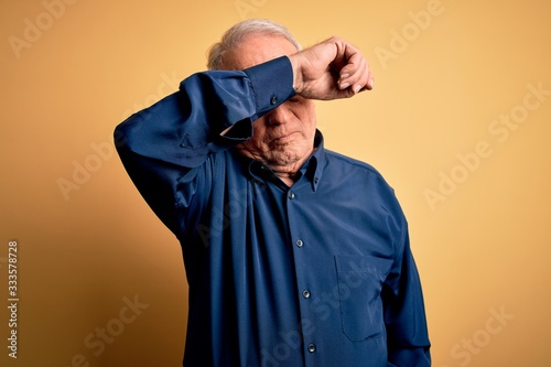 Grey haired senior man wearing casual blue shirt standing over yellow background covering eyes with arm, looking serious and sad. Sightless, hiding and rejection concept © Krakenimages.com