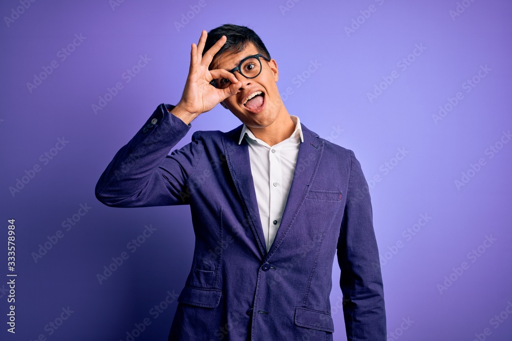 Young handsome business man wearing jacket and glasses over isolated purple background doing ok gesture with hand smiling, eye looking through fingers with happy face.